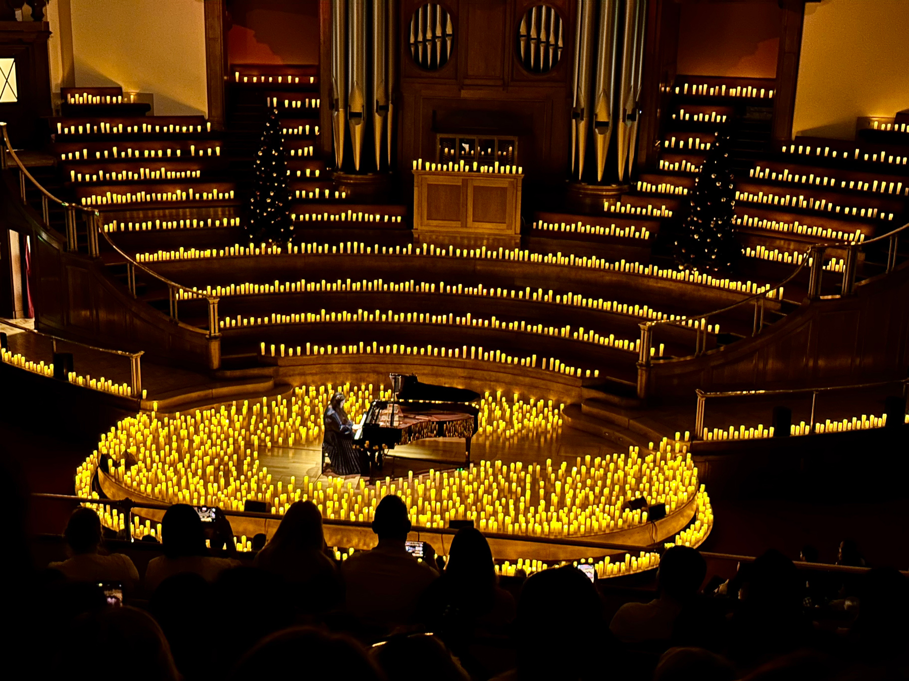 A view of a round stage covered in candles, with a grand piano in the center. Framed by a large pipe organ and candles covering the stepped choir seating.