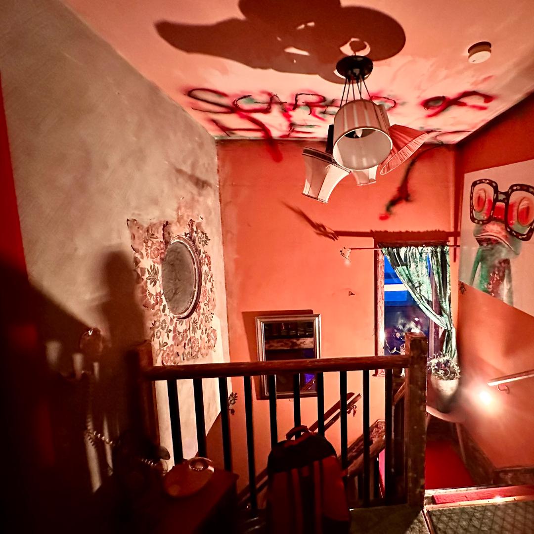 A landing bathed in red light, several paintings are on the walls and the ceiling is daubed with the message 'Scared of heights'