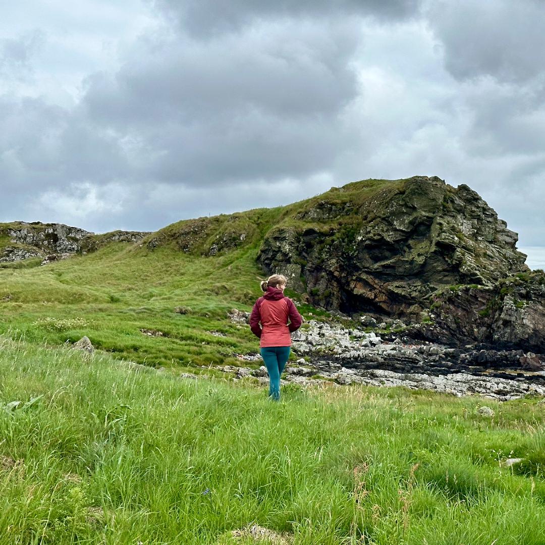 Charlotte walking along a grassy coastline with a large rocky outcrop in the background