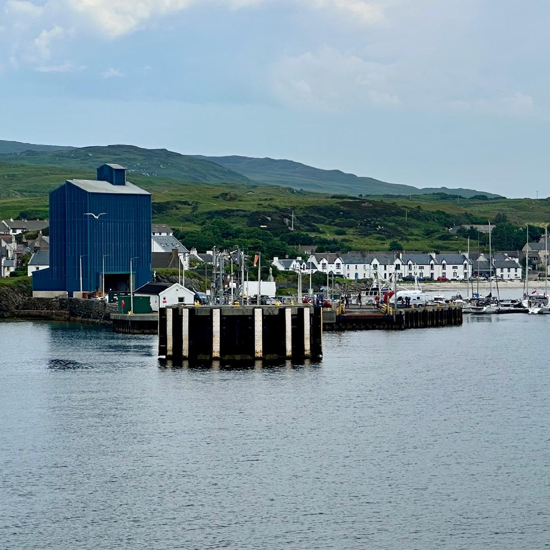 Port Ellen viewed from the ferry on the way in. A thin strip of white houses lines the bay where fishing boats are moored, green hills tower behind the town