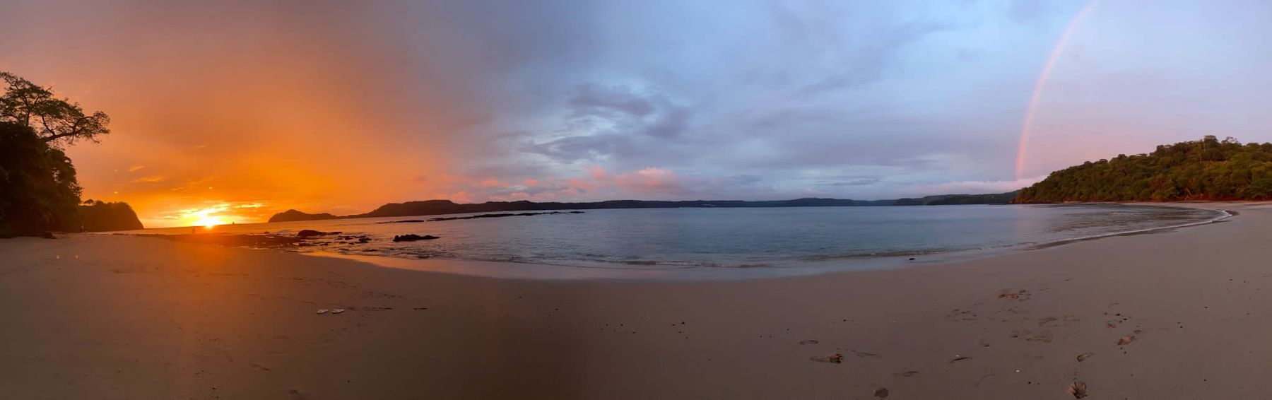 A panorama of a beach with an orange sunset on one side and a large rainbow on the other